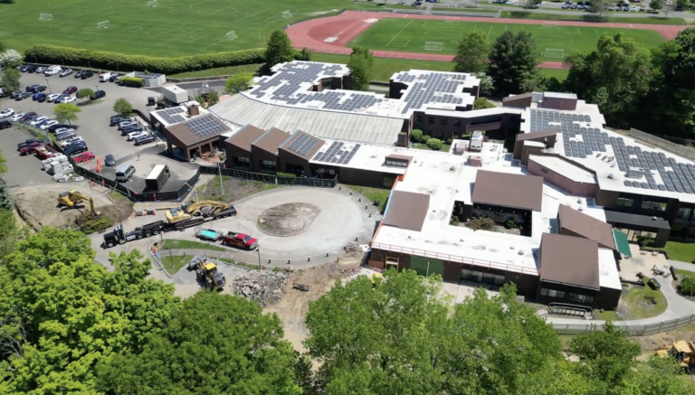 Aerial view of a large campus building with extensive solar panels on the roof, a construction area nearby, and a sports field in the background.